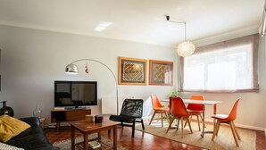 A modern living room with a flat-screen TV, a black leather sofa, a wooden coffee table, and a dining area with orange chairs. - Hipster´s dream by the river (Porto)