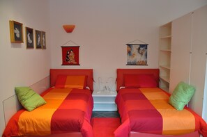 Quarto com duas camas, cada uma com roupa de cama em vermelho e laranja, um criado-mudo branco e um painel de parede vermelho.