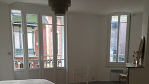 Une chambre à coucher avec un lit, un plancher en bois, un bureau avec une chaise et de grandes fenêtres donnant sur des immeubles.