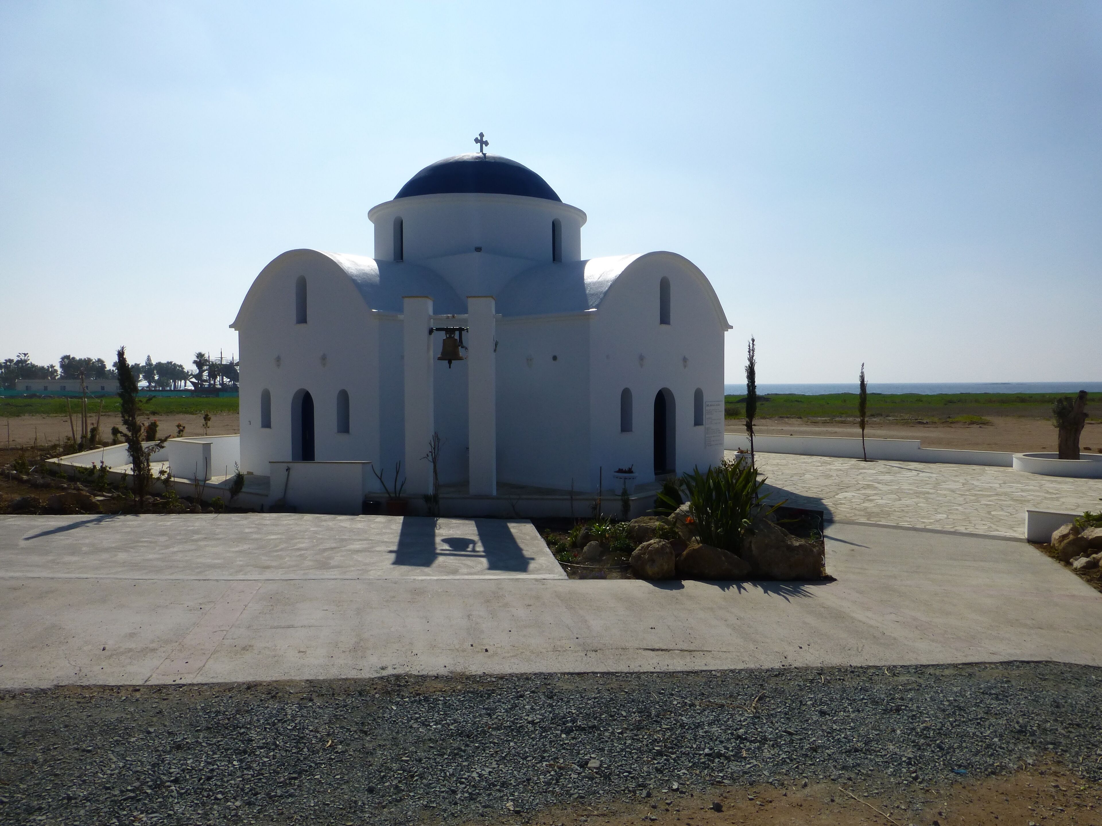 A white church with a blue dome and a bell tower, surrounded by a paved area and greenery.