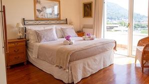 Ein Schlafzimmer mit einem großen Bett, einem Nachttisch, einem Sessel und Blick ins Freie.
