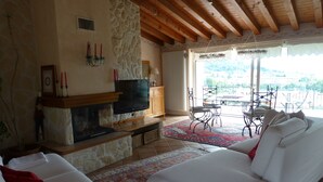 Ein Wohnzimmer mit Kamin, weißen Sofas, einem Fernseher und Blick ins Freie.