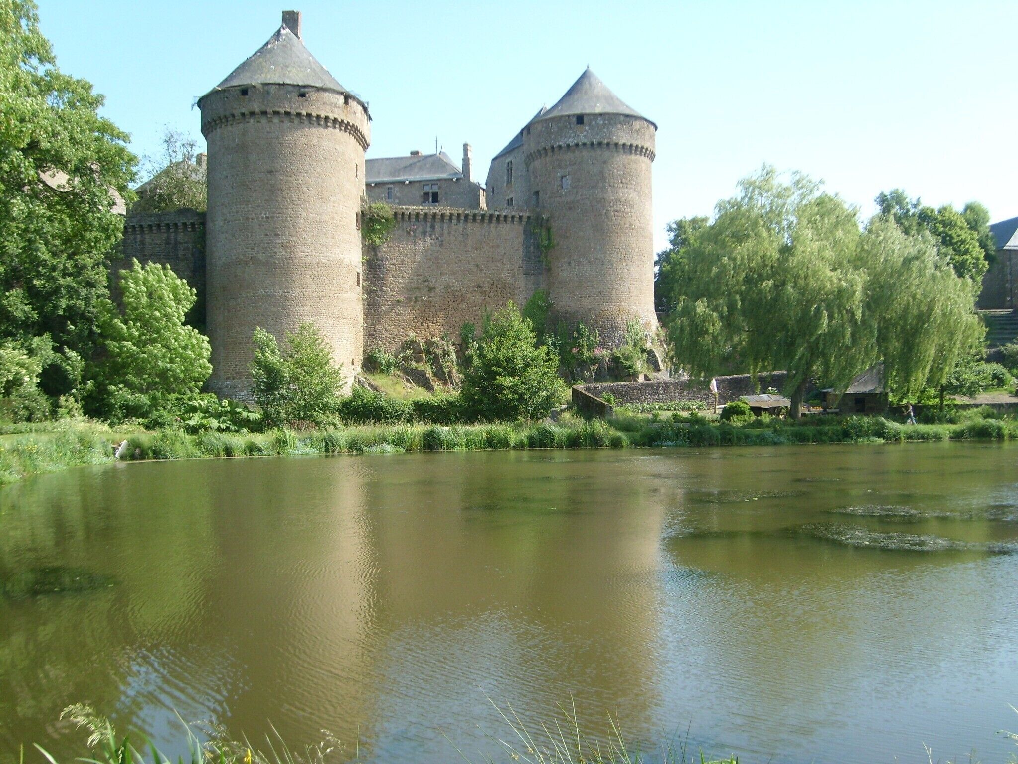 Een historisch kasteel met twee torentjes, omringd door weelderige begroeiing en een rustige rivier.
