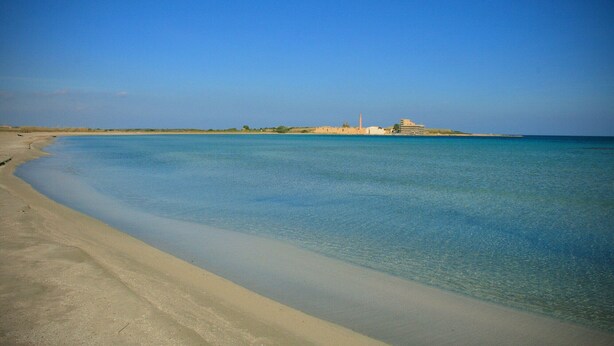 A beach with clear blue water, a sandy shore, and a distant building with a tower.