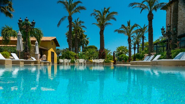 Una piscina blava i nítida amb gandules i ombrel·les, envoltada de palmeres i un edifici amb teulada de rajola.