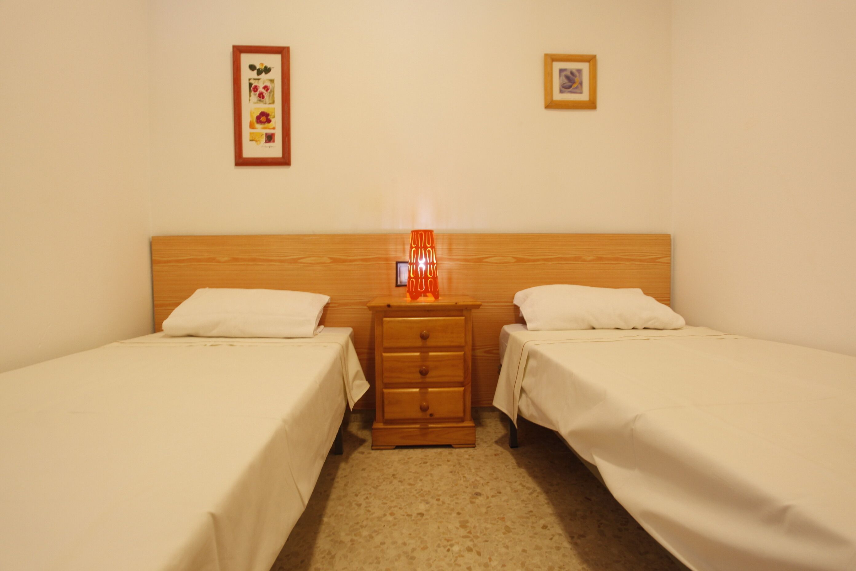 Ein Zimmer mit zwei Einzelbetten, einem Nachttisch aus Holz mit Lampe und zwei gerahmten Bildern an der Wand.