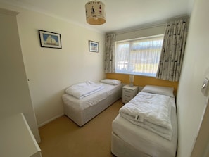Een slaapkamer met twee eenpersoonsbedden, een nachtkastje met een lamp, een raam met gordijnen en twee ingelijste foto's aan de muur.