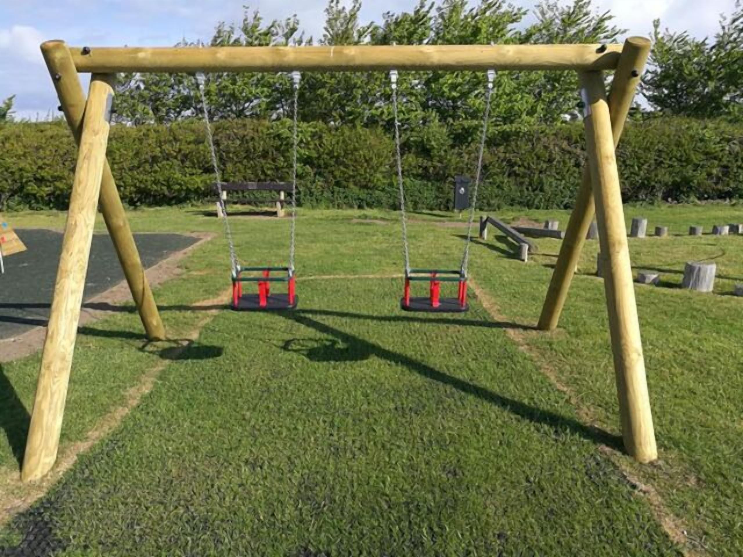 A wooden swing set with two swings, set on a grassy area with trees in the background.