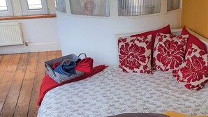 Un dormitorio con una cama, almohadas estampadas en rojo y blanco, una maleta gris y una manta roja en el suelo.