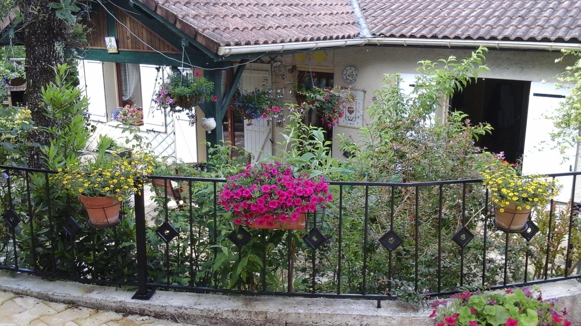 Una casa con techo de tejas, balcón con barandilla metálica y varios maceteros con plantas y flores.