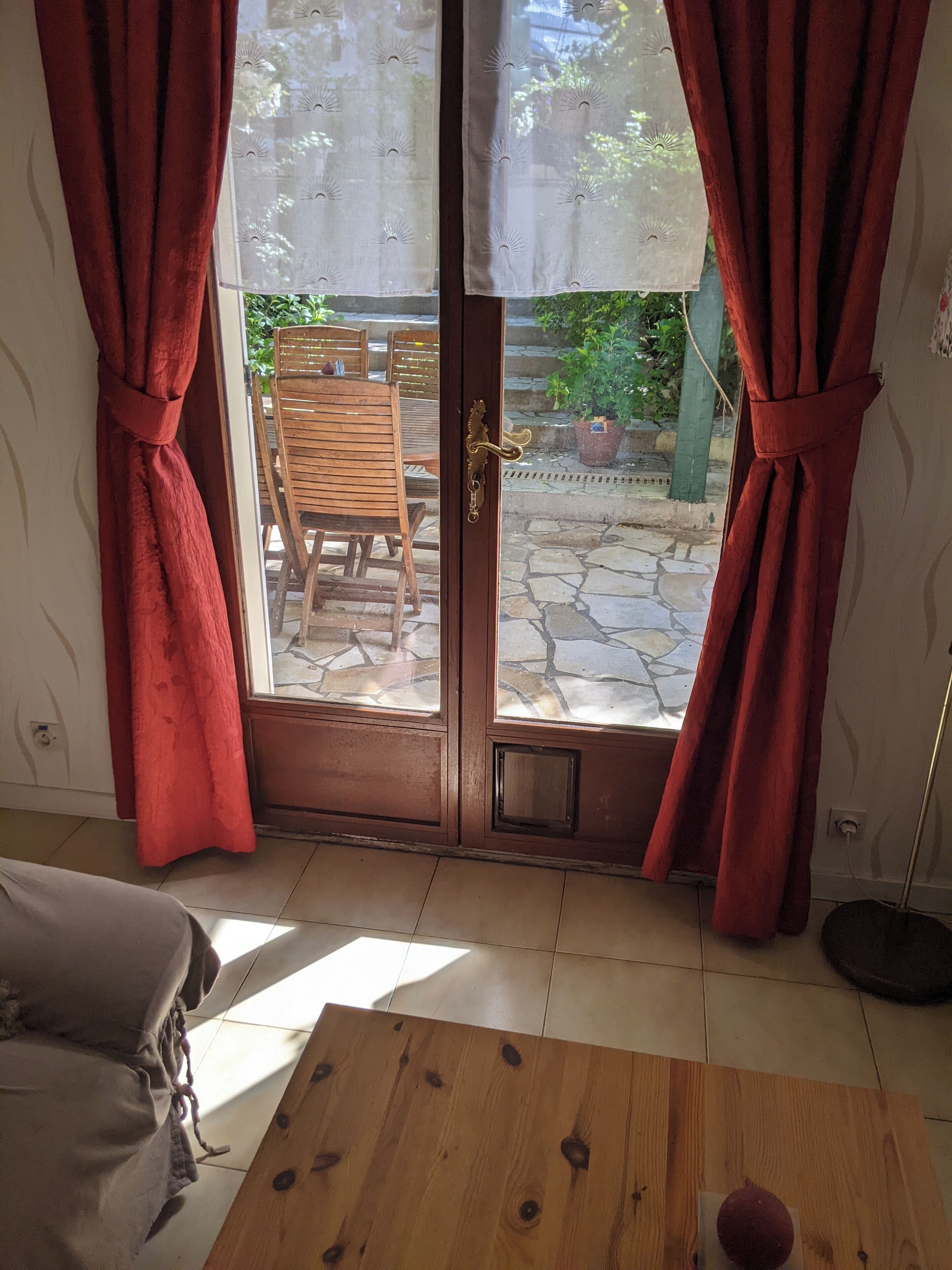 Ein Zimmer mit Holztür, roten Vorhängen, einem Holztisch und Blick auf eine Terrasse mit Stuhl und Pflanzen.