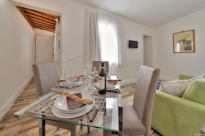 Uma área de refeições com uma mesa de vidro posta para dois, uma garrafa de vinho e um sofá verde ao fundo.