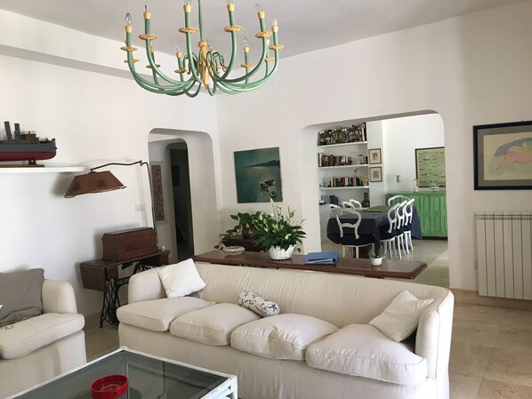 A living room with a green chandelier, a white sofa, a dining area with a green table, and a kitchen visible through an archway.