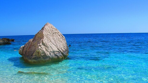 Mar azul transparente con una roca grande al frente y una playa arenosa.