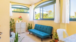 Una habitación acogedora con un sofá azul, una silla blanca y una pequeña mesa con un libro. Hay plantas en macetas y un mueble con un televisor.