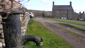 Un paysage rural avec un mur de pierres, une voiture blanche garée sur un chemin de terre et un grand bâtiment en pierre doté de nombreuses fenêtres.