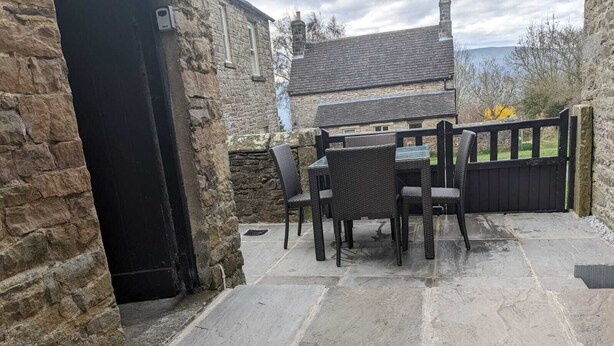 Une terrasse en pierre avec une table et des chaises, un mur en pierre et, au loin, une maison.