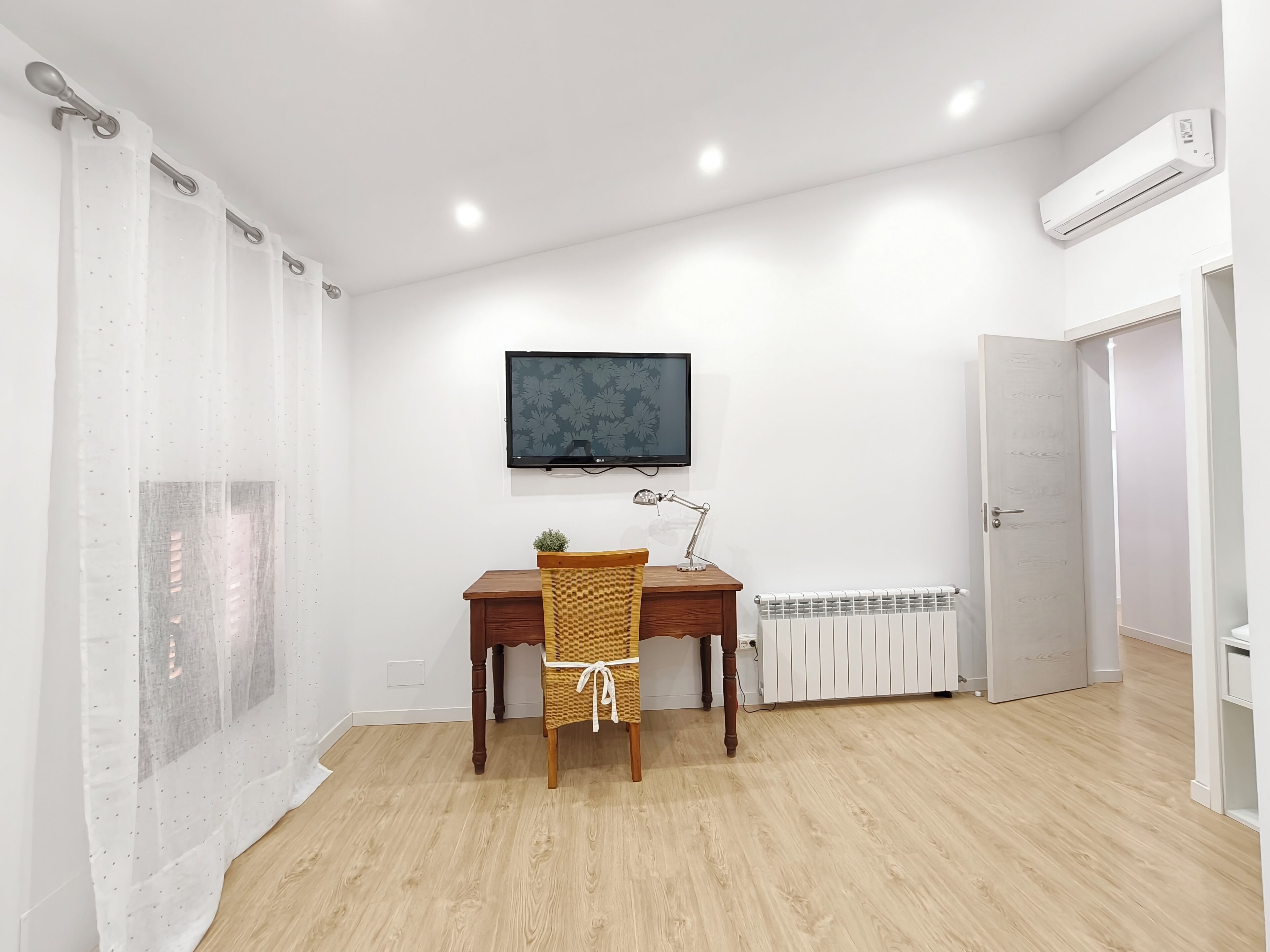 A room with a wooden desk, a chair, a television mounted on the wall, a radiator, and a door.