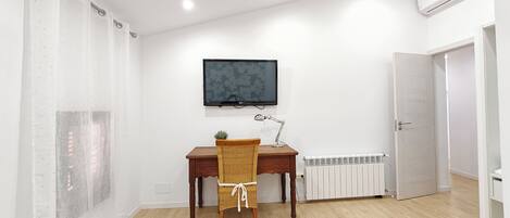 A room with a wooden desk, a chair, a television mounted on the wall, a radiator, and a door.
