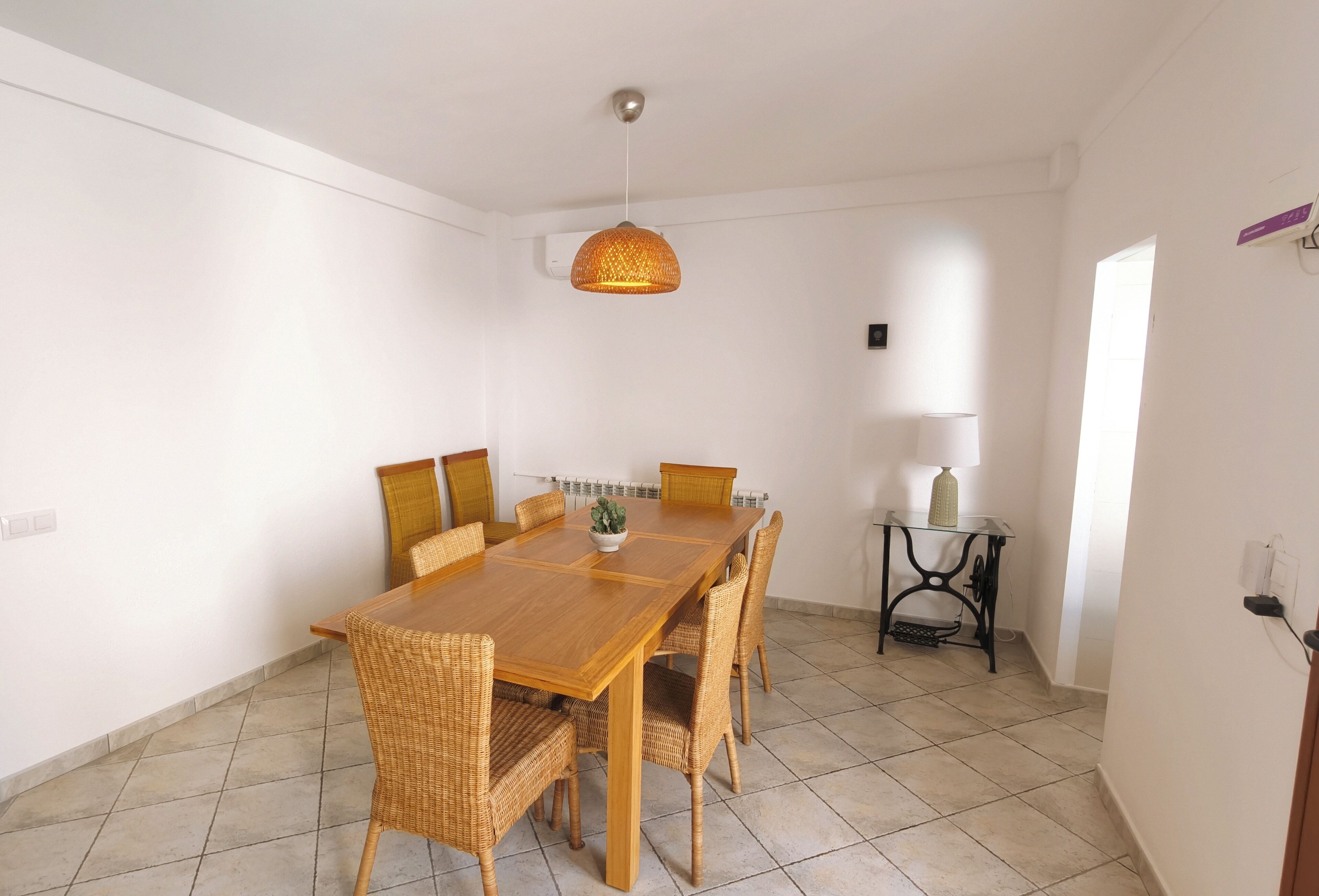 A dining area with a wooden table and wicker chairs, a hanging light fixture, and a small table with a lamp.
