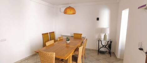 A dining area with a wooden table and wicker chairs, a hanging light fixture, and a small table with a lamp.