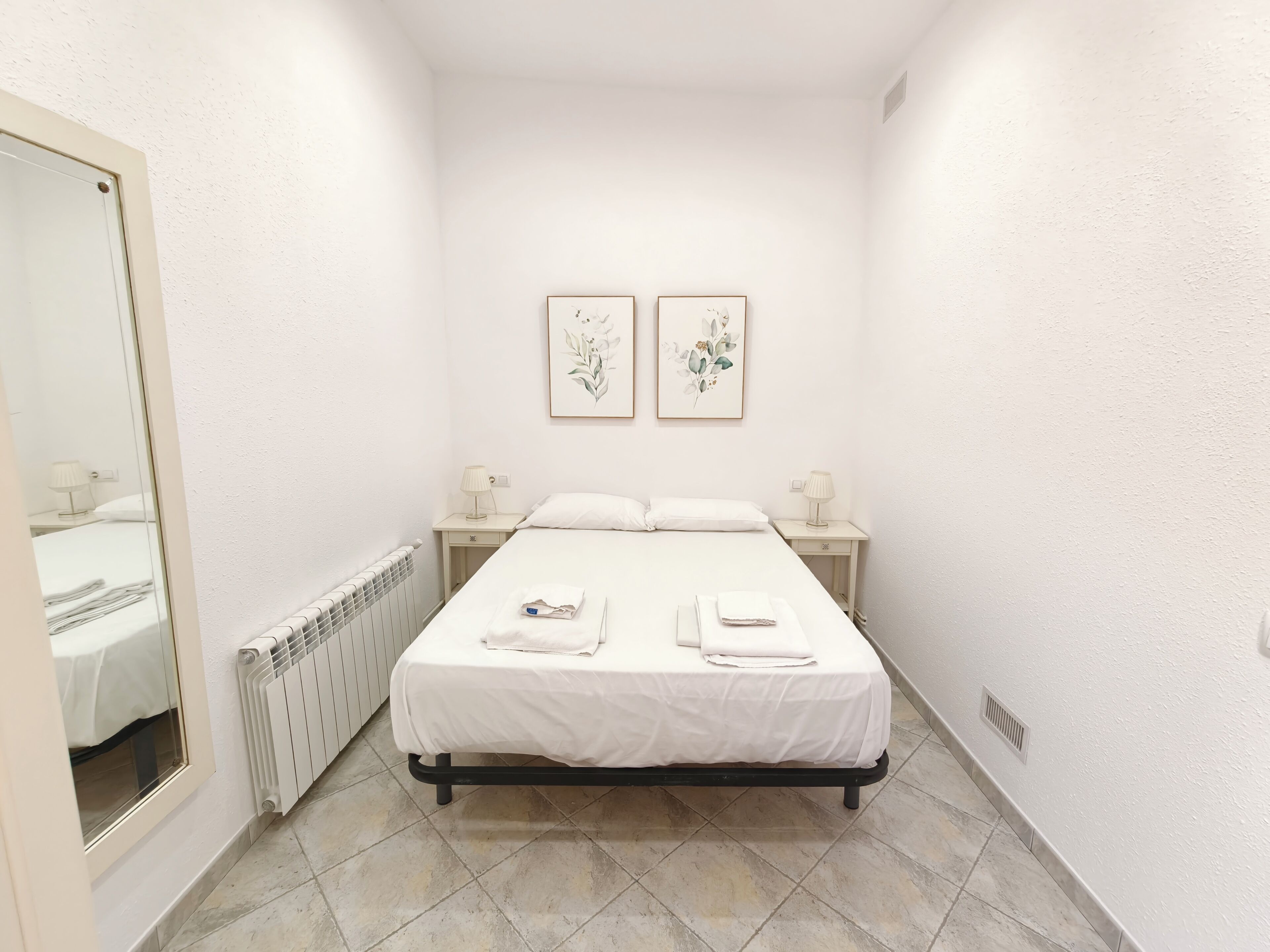 Une petite chambre propre, avec un lit, deux tables de chevet dotées de lampes, un radiateur et deux œuvres d’art florales encadrées accrochées au mur.