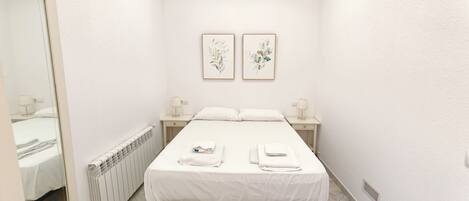 Une petite chambre propre, avec un lit, deux tables de chevet dotées de lampes, un radiateur et deux œuvres d’art florales encadrées accrochées au mur.