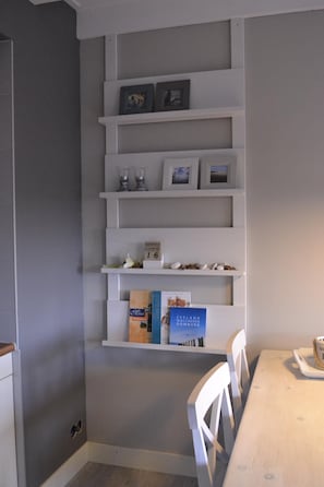 Quarto com parede branca, prateleira embutida com objetos decorativos, mesa de madeira e cadeiras brancas.