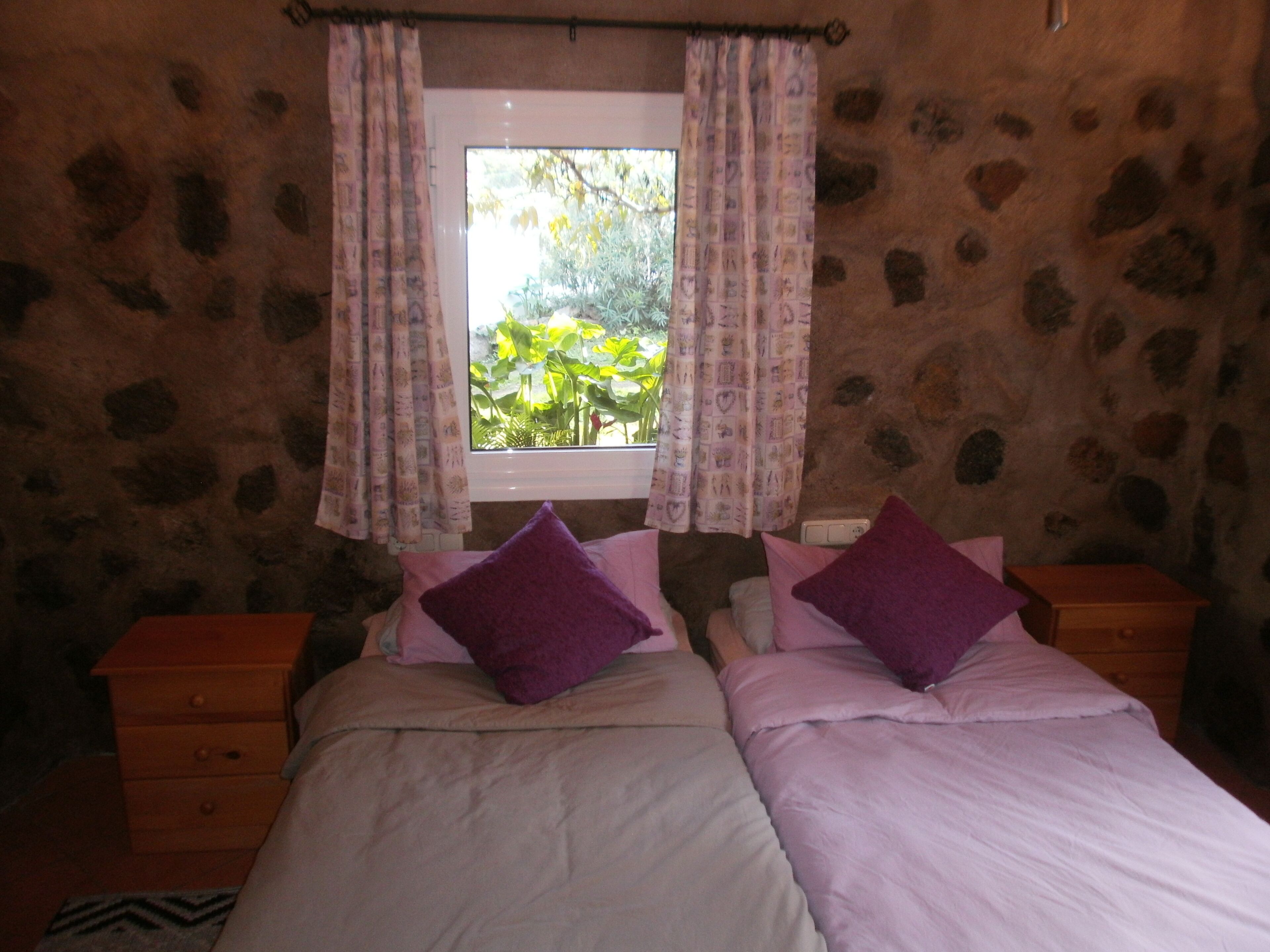 A bedroom with two beds, a window with patterned curtains, and a stone wall.