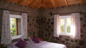 A bedroom with a stone wall, wooden ceiling beams, two beds with purple pillows, and windows with patterned curtains. - Traditional Canarian Finca in Las Lajas, Icod de los Vinos, 4 persons (Icod de los Vinos)