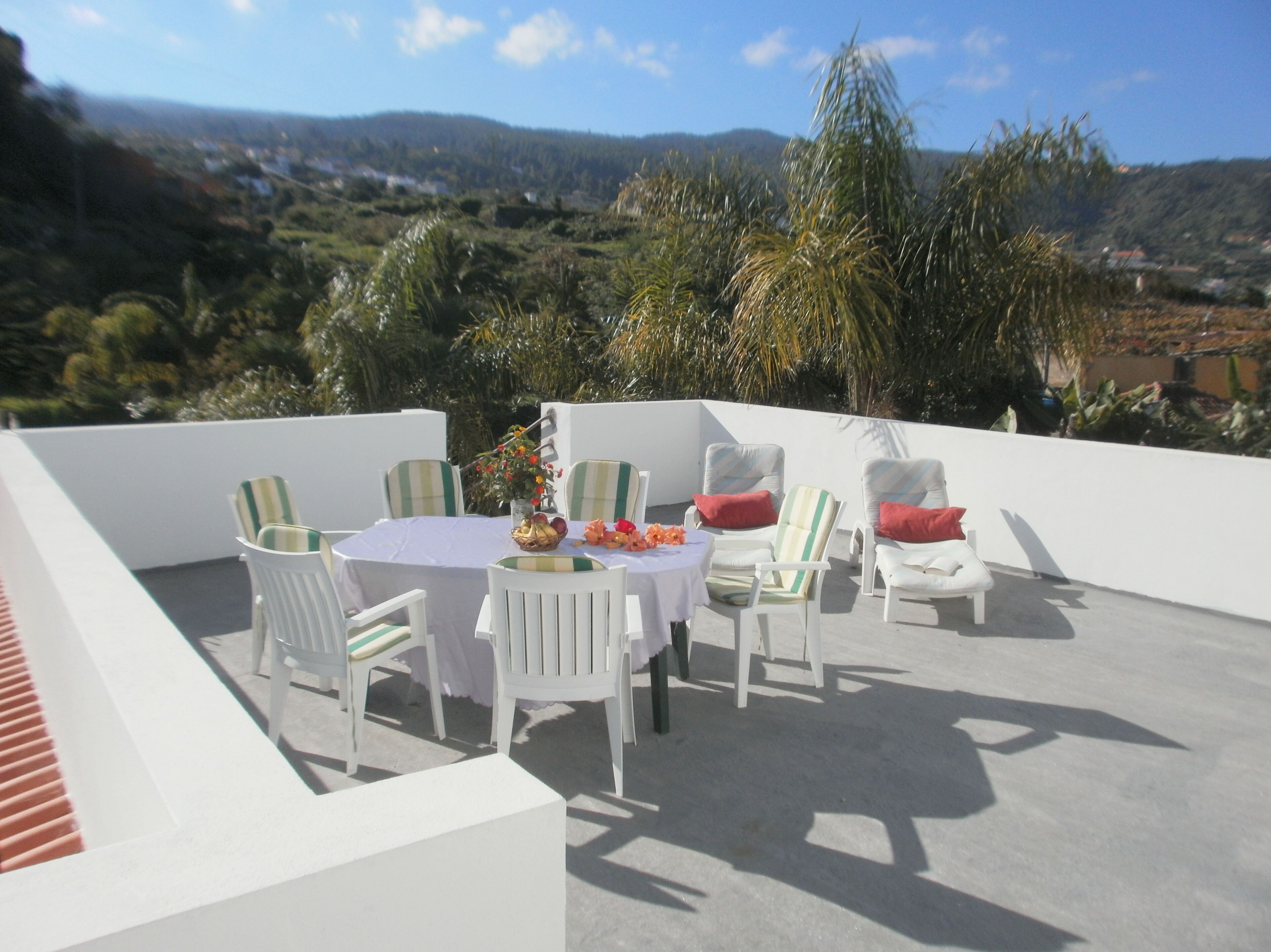 Um terraço com uma mesa redonda e cadeiras, uma pequena mesa com uma cesta de frutas, e vista para uma paisagem acidentada.