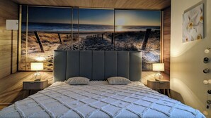 Une chambre à coucher avec un grand lit, deux tables de chevet et une grande œuvre d’art accrochée au mur, représentant une scène de plage.
