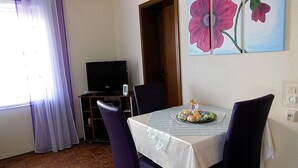 Zona pranzo con tavolo apparecchiato per due, televisione su un mobile e un quadro con fiori sulla parete.