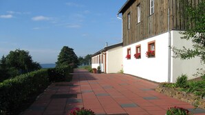 A pathway leading to a building with a white facade and wooden upper section, flanked by greenery and potted plants.