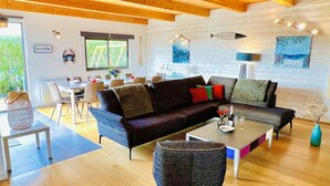 Een ruime woonkamer met een houten plafond, een donkergroene bank, een klein rond tafeltje en een eethoek met stoelen.