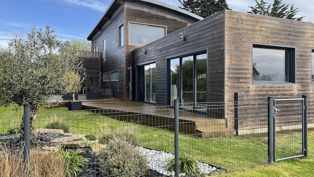 Een modern huis met twee verdiepingen, met een houten gevel, grote ramen en een verzorgde tuin met een hek.