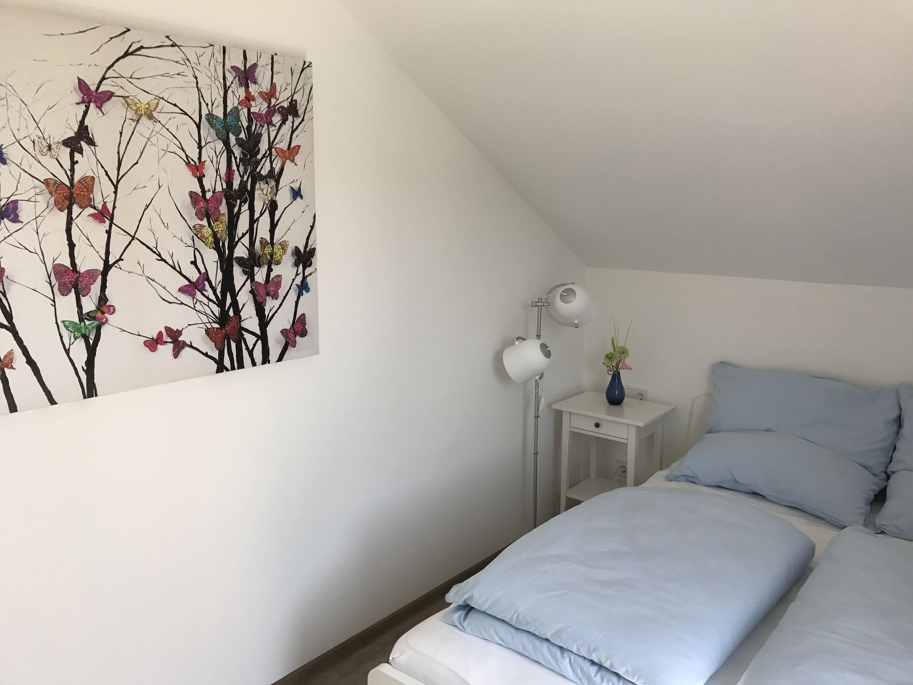 A bedroom with a bed, a nightstand, a lamp, and a wall art of butterflies on branches.