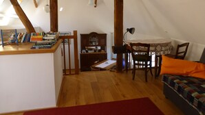 A cozy attic room with wooden beams, a dining table with chairs, a small bookshelf, and a red rug.