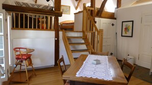 A wooden dining table with a lace tablecloth, surrounded by wooden chairs. A staircase with wooden railings leads to an upper level.