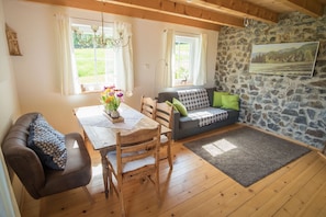 A cozy room with a stone wall, wooden floor, a dining table with chairs, a sofa with cushions, and a window with curtains.