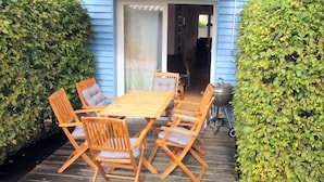 Une terrasse extérieure avec une table et des chaises en bois, un barbecue et une maison bleue en arrière-plan.