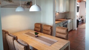 Un coin repas avec une table en bois, des chaises en cuir et des luminaires suspendus. On aperçoit une cuisine à l’arri ère-plan, avec des meubles blancs et un évier.