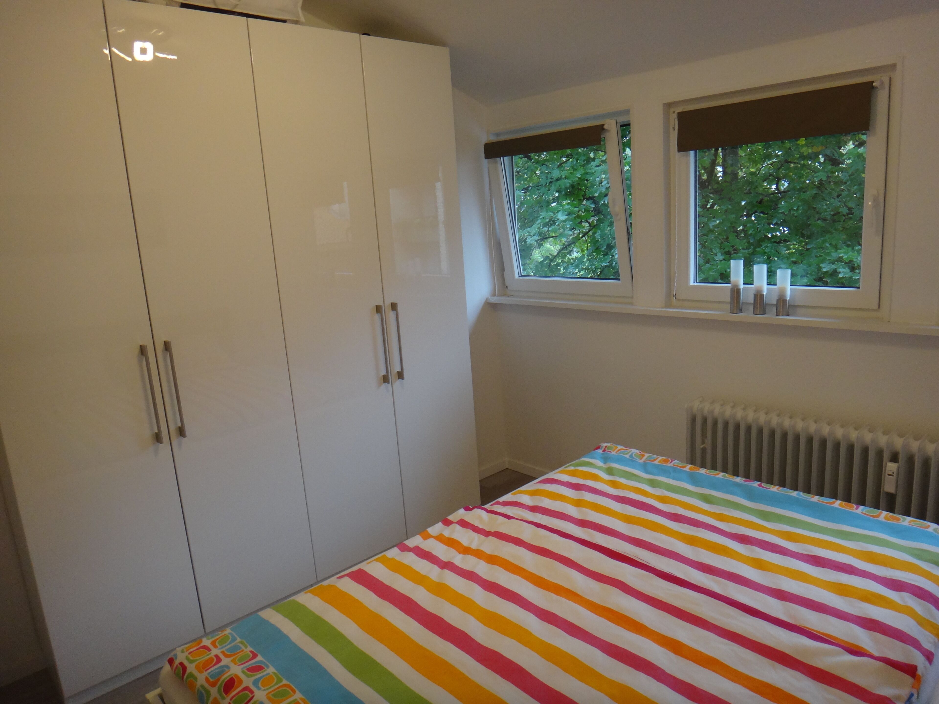 A bedroom with a striped bedspread, a white wardrobe, and a window with a view of trees.