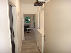 A narrow hallway with a wooden floor, a small table, and a bench. There is a door on the right and a window at the end of the hallway.