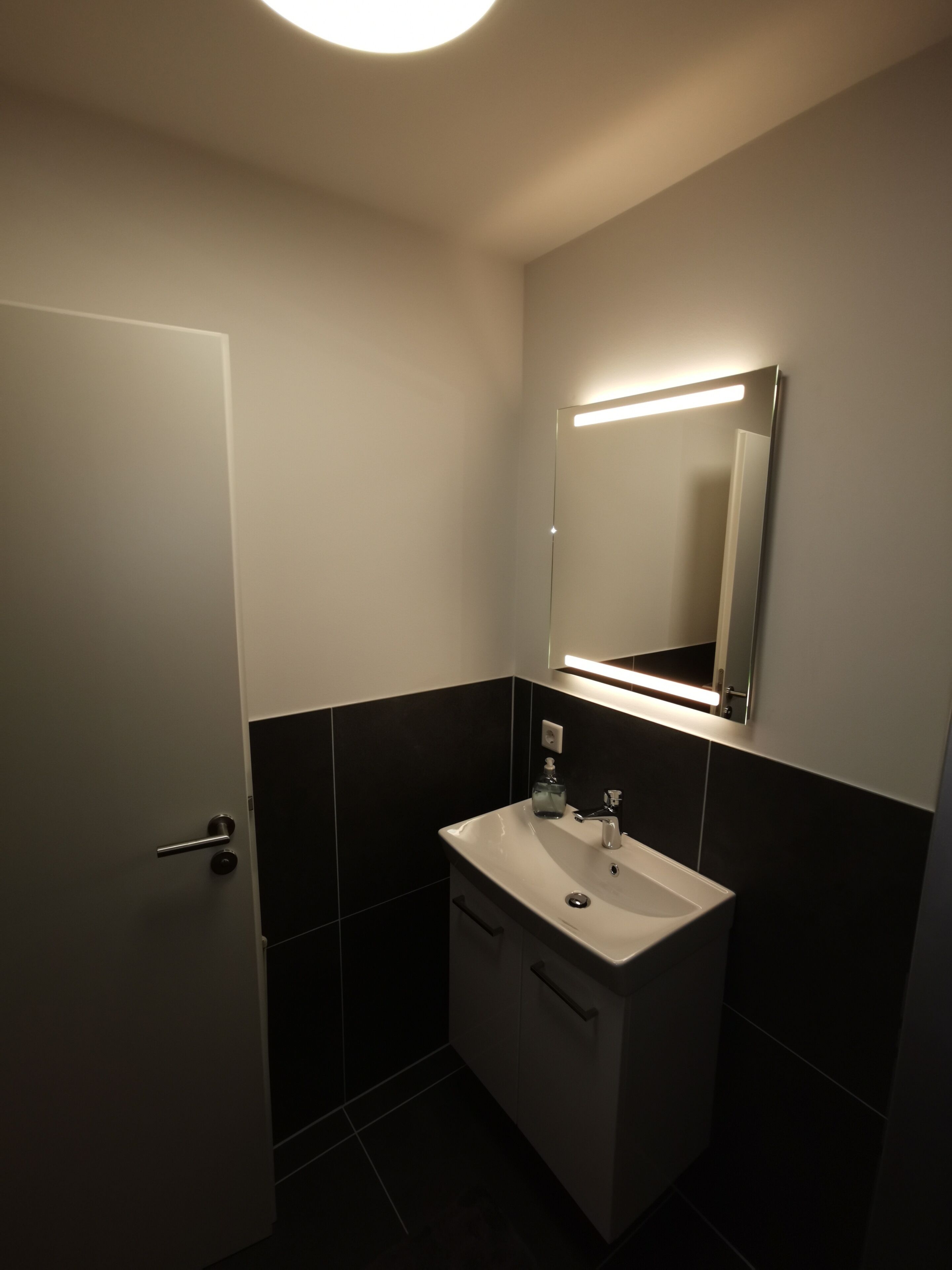 A bathroom with a white sink, dark tiled walls, a mirror above the sink, and a door to the left.