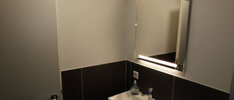 A bathroom with a white sink, dark tiled walls, a mirror above the sink, and a door to the left.