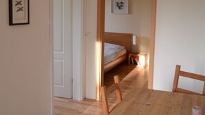 A room with a wooden dining table and chairs, a bed with a white headboard, and a door leading to another room. - Pferdehof Muhlitz on Rügen, an idyll for individualists (Samtens)