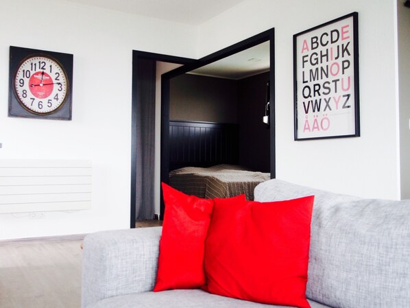 Une pièce avec un canapé gris, un coussin rouge, une horloge et une affiche illustrant l’alphabet, encadrée et accrochée au mur.