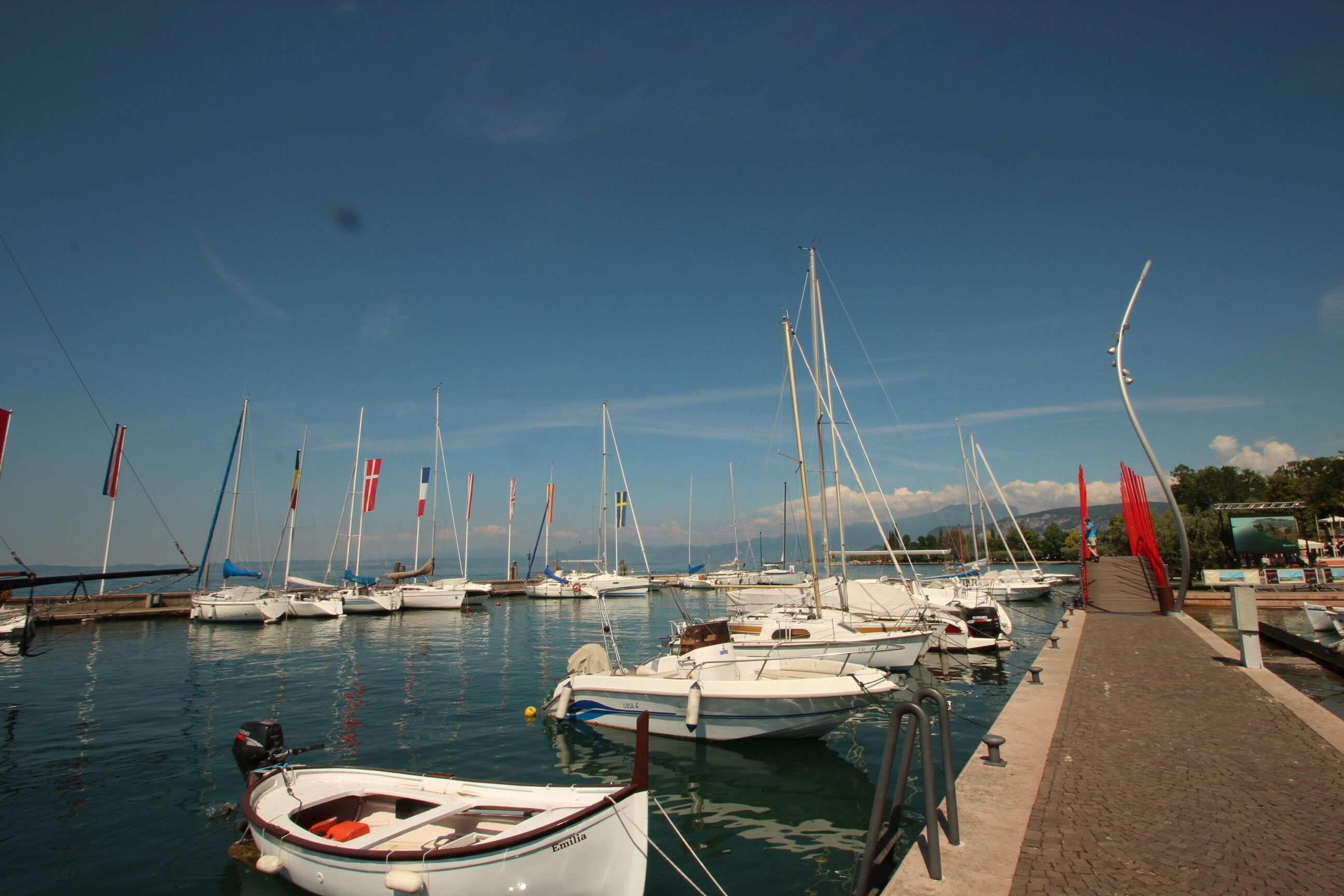 A marina with multiple boats docked, a clear sky, and a paved walkway along the water.