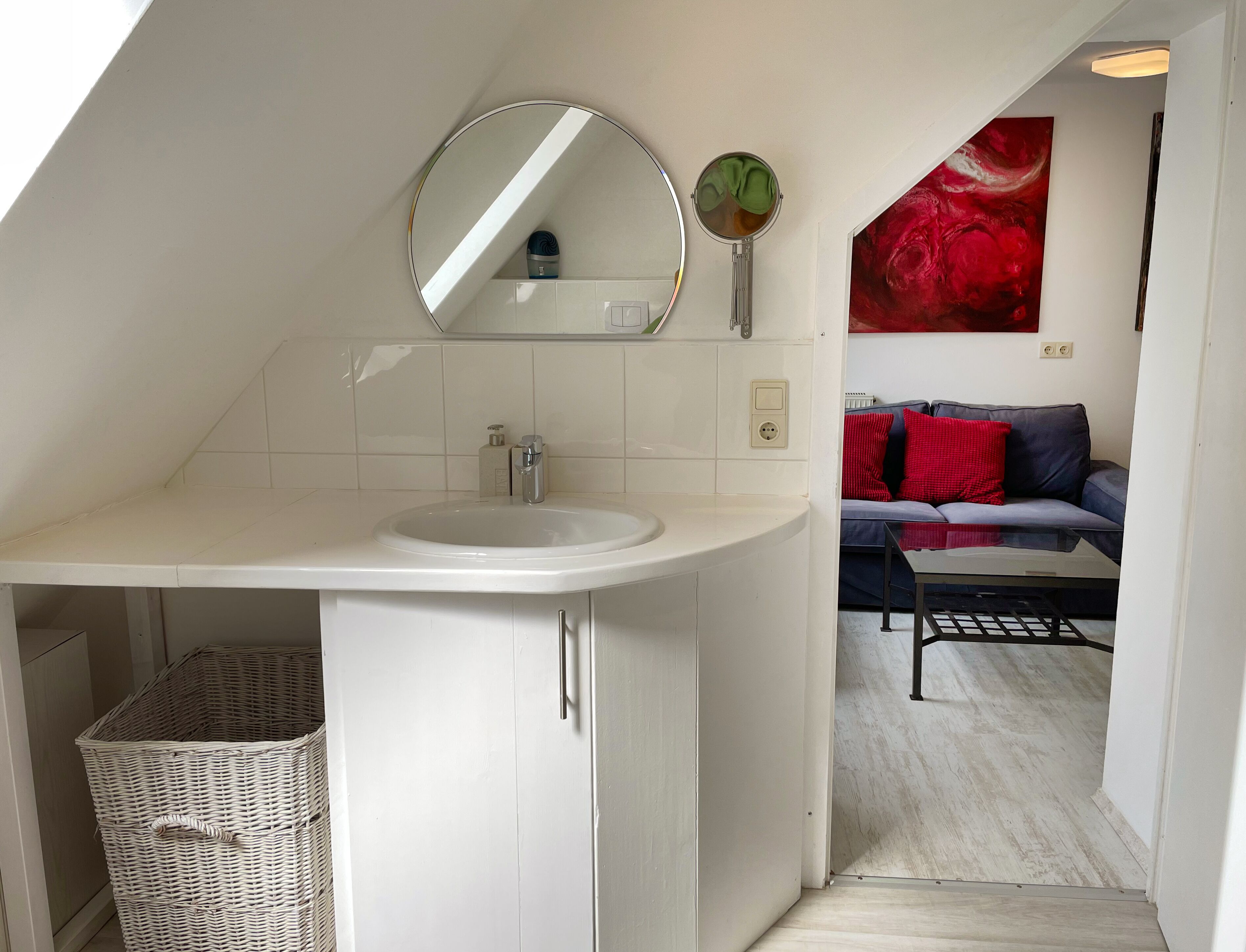 A modern bathroom with a white sink, a round mirror, and a small round mirror on the wall. There is a wicker basket under the sink and a red abstract painting on the wall.